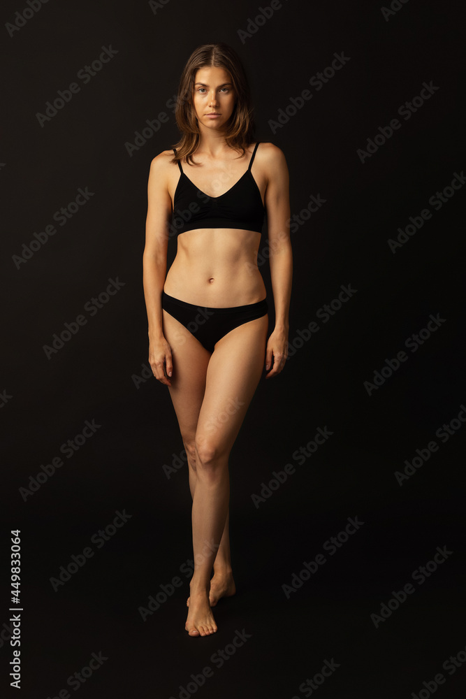 Portrait of young beautiful slim tanned woman in black lingerie posing isolated over dark studio background. Natural beauty, wellness, healthy lifestyle concept.
