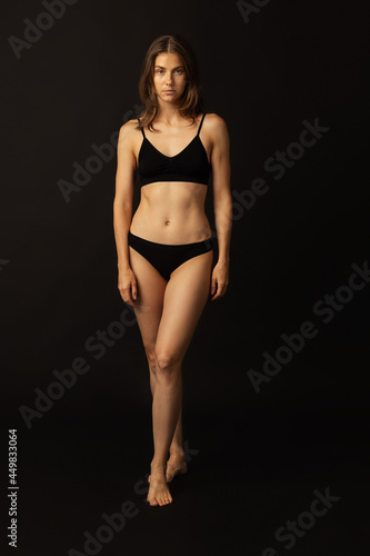 Portrait of young beautiful slim tanned woman in black lingerie posing isolated over dark studio background. Natural beauty, wellness, healthy lifestyle concept.