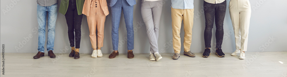 Cropped image of legs of people standing in a row near the wall waiting for an interview. Unidentified men and women in business and casual clothes and shoes stand leaning against a light wall.