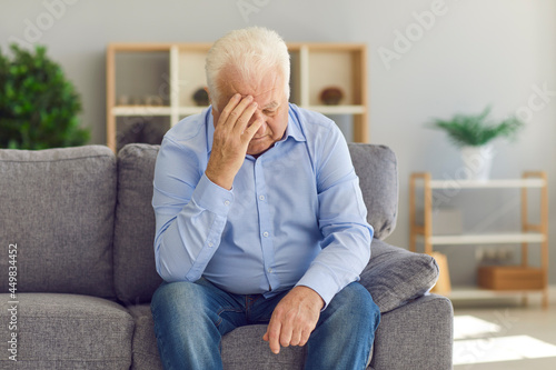Retired old granddad sitting on couch at home, crying, grieving over loss of wife, feeling depressed and lonely. White-haired senior man touching forehead suffering from severe bad headache