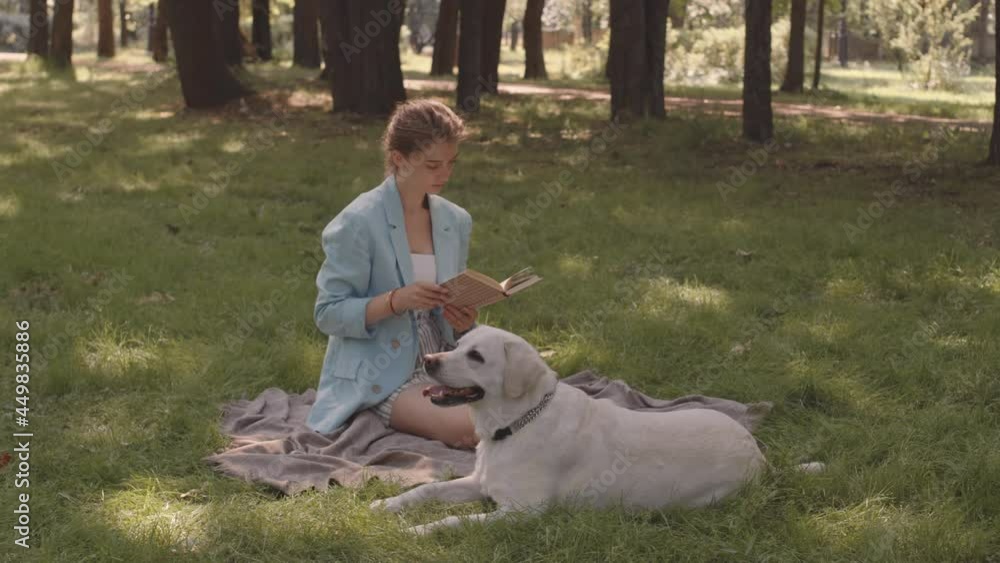 Long arc of female Caucasian dog owner reading book in forest, sitting on lawn next to her Labrador Retriever pet Long arc of female Caucasian dog owner reading book in forest, sitting on lawn next to her Labrador Retriever pet