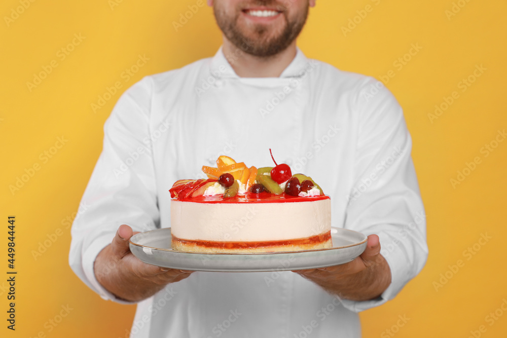 Happy professional confectioner in uniform holding delicious cake on yellow background, closeup