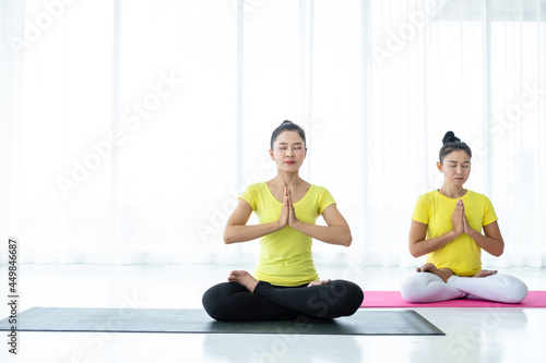 Two young Asian women workout practicing yoga in yellow dress or pose with a trainer and practice meditation wellness lifestyle and health fitness concept in a gym.