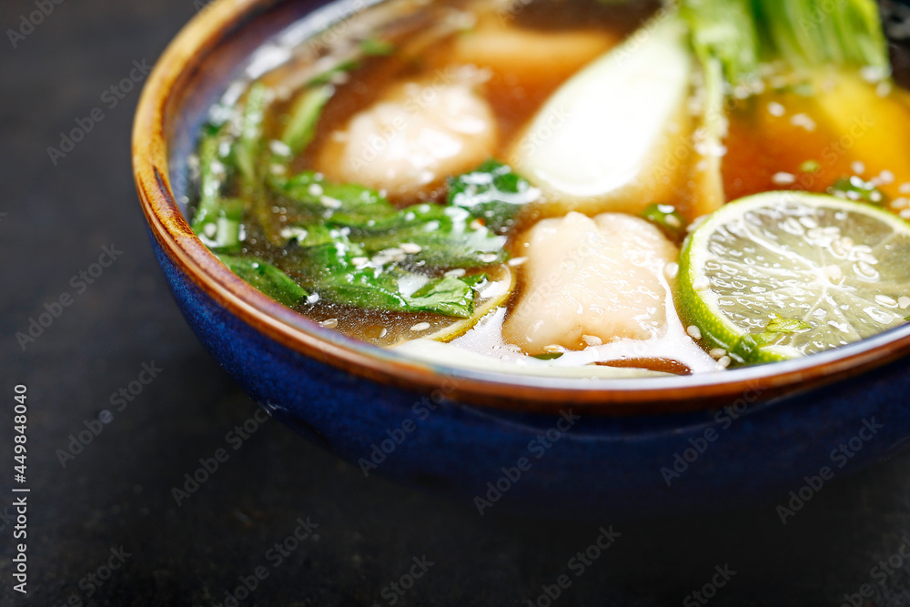 A close-up of oriental soup. Vegetable and meat broth with fresh herbs, sesame, lime, shrimp, vegetable dumplings in an oriental bowl, on black stony background. A delicacy of Eastern cuisine.