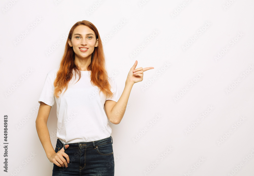 Smiling young red-haired woman isolated on gray studio background pointing with finger at blank copy space aside Smiling young red-haired woman isolated on gray studio background pointing with finger at blank copy space aside