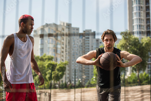Man with basketball ball looking at camera near african american sportsman outdoors