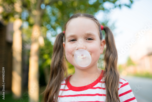 Fotografie Portrait of smiling girl with two ponytails looking at camera and inflating bubble with chewing gum outdoor
