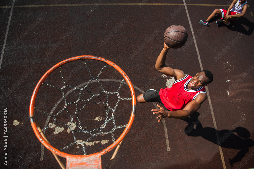 Fototapeta premium Top view of excited african american man with basketball ball jumping near hoop