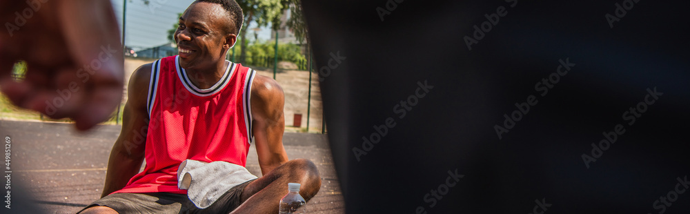 Positive african american sportsman with towel sitting near water on playground, banner Positive african american sportsman with towel sitting near water on playground, banner