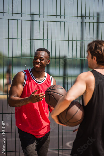 Happy african american sportsman holding basketball ball near fence and blurred friend