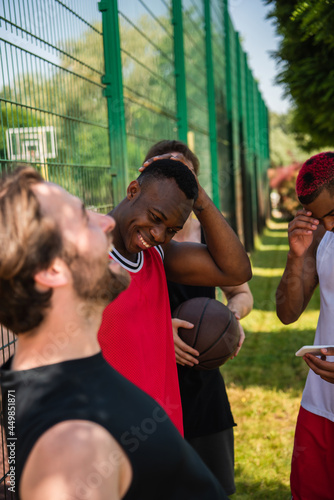 Fototapeta Naklejka Na Ścianę i Meble -  Interracial basketball players with ball and smartphone laughing outdoors