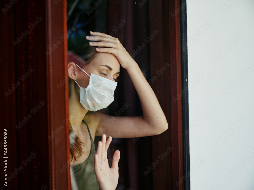 woman in medical mask holds hand on forehead quarantine view from window