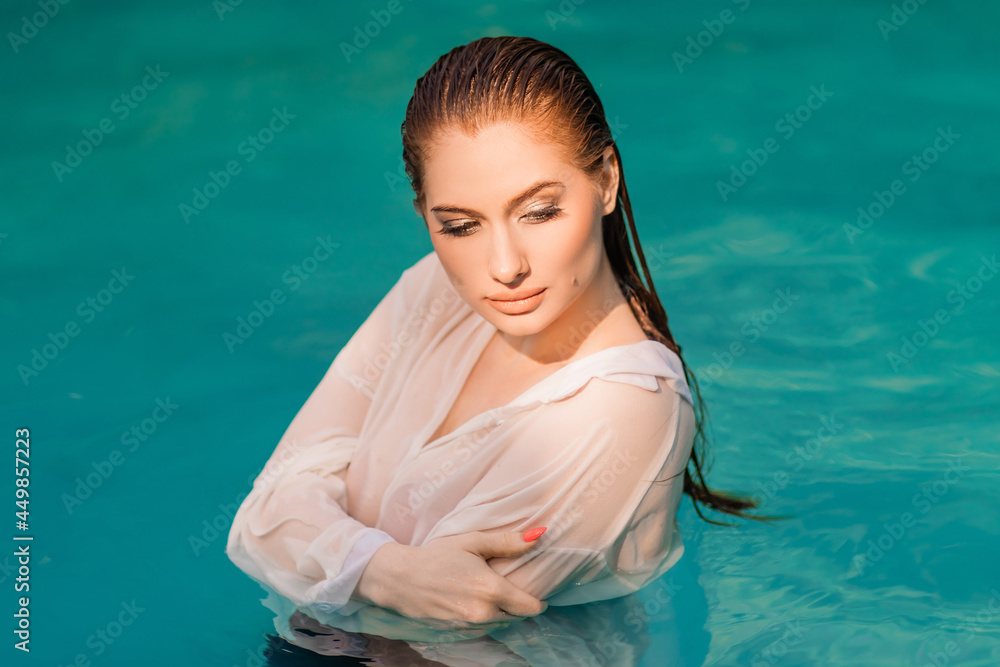 Portrait of a beautiful girl with wet hair dressed in a white shirt standing up to her chest in the pool.