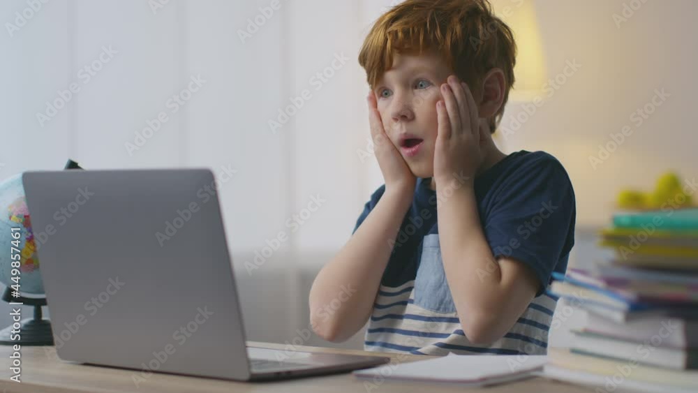 Portrait of shocked little redhead boy looking at laptop with amazed expression, touching his cheeks in amazement Portrait of shocked little redhead boy looking at laptop with amazed expression, touching his cheeks in amazement