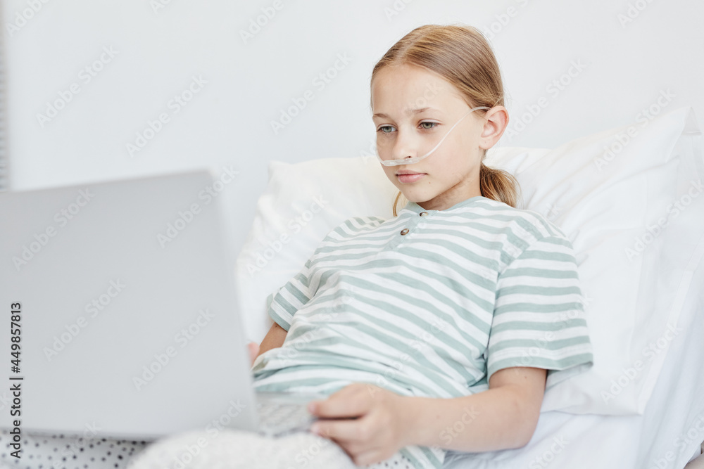 Minimal portrait of little girl laying in hospital bed and using laptop with oxygen support, copy space Minimal portrait of little girl laying in hospital bed and using laptop with oxygen support, copy space