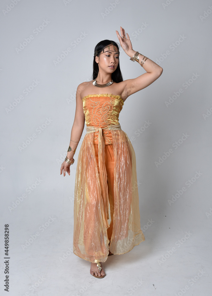 Full length portrait of pretty young asian woman wearing golden Arabian robes like a genie, standing pose isolated on studio background. Full length portrait of pretty young asian woman wearing golden Arabian robes like a genie, standing pose isolated on studio background.