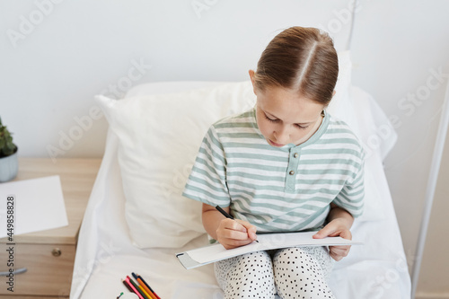 Minimal portrait of little girl in hospital room drawing pictures while sitting on bed, copy space