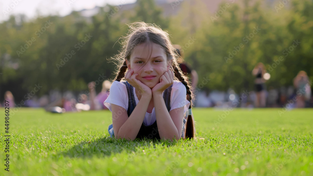 Smiling cute girl with braids lying on green lawn in park looking at camera