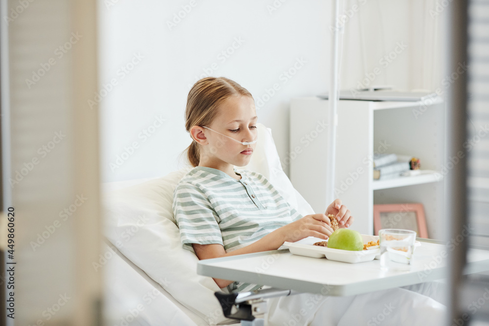 Minimal side view portrait of girl eating healthy lunch while lying on bed in hospital room