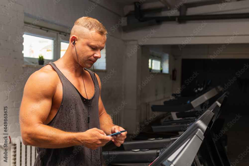 Man in gym using smartphone with headphones sportsman in headphones choose music on his smartphone at gym. Man in gym using smartphone with headphones sportsman in headphones choose music on his smartphone at gym.