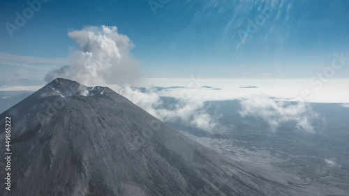 A conical stratovolcano rises above the clouds. Fumaroles are visible above the crater. The rays of the sun in the blue sky. Aerial view. Kamchatka