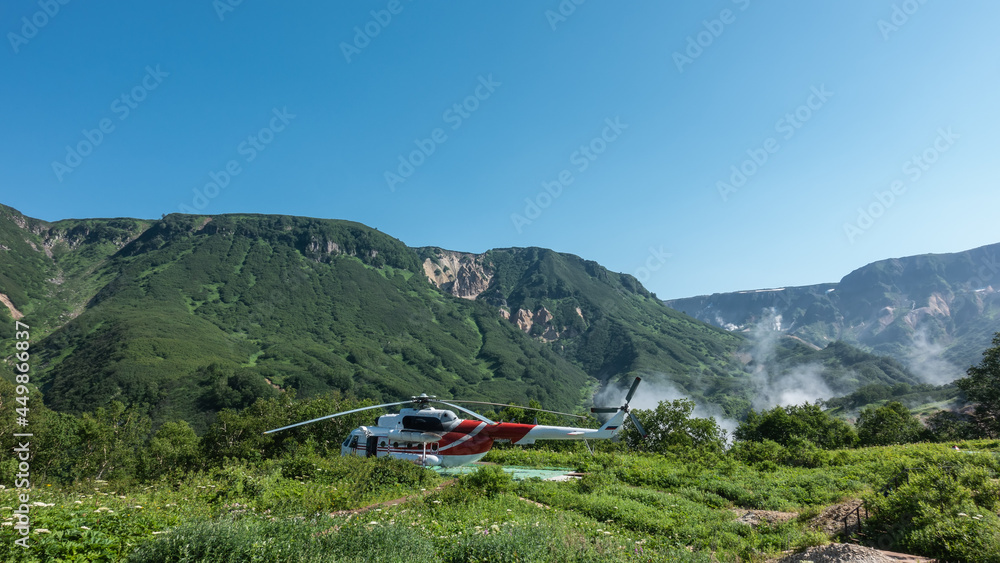 The helicopter is standing on the landing pad. There is lush grass and wildflowers in the meadow. There is green vegetation on the mountain slopes. Steam rising from the Valley of Geysers is visible. The helicopter is standing on the landing pad. There is lush grass and wildflowers in the meadow. There is green vegetation on the mountain slopes. Steam rising from the Valley of Geysers is visible.