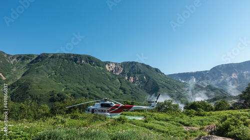 The helicopter is standing on the landing pad. There is lush grass and wildflowers in the meadow. There is green vegetation on the mountain slopes. Steam rising from the Valley of Geysers is visible.