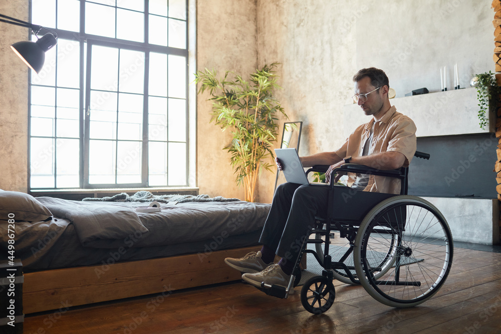 Full length portrait of modern young man in wheelchair using laptop at home in designer interior, copy space