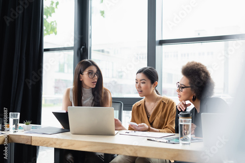 multiethnic businesswomen talking near laptop in office on blurred foreground