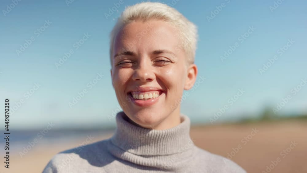 Head and shoulders fade in closeup portrait of happy young woman with blonde short hair and in gray turtleneck sweater looking at camera and laughing on beach. Travel, happiness and freedom concepts Head and shoulders fade in closeup portrait of happy young woman with blonde short hair and in gray turtleneck sweater looking at camera and laughing on beach. Travel, happiness and freedom concepts