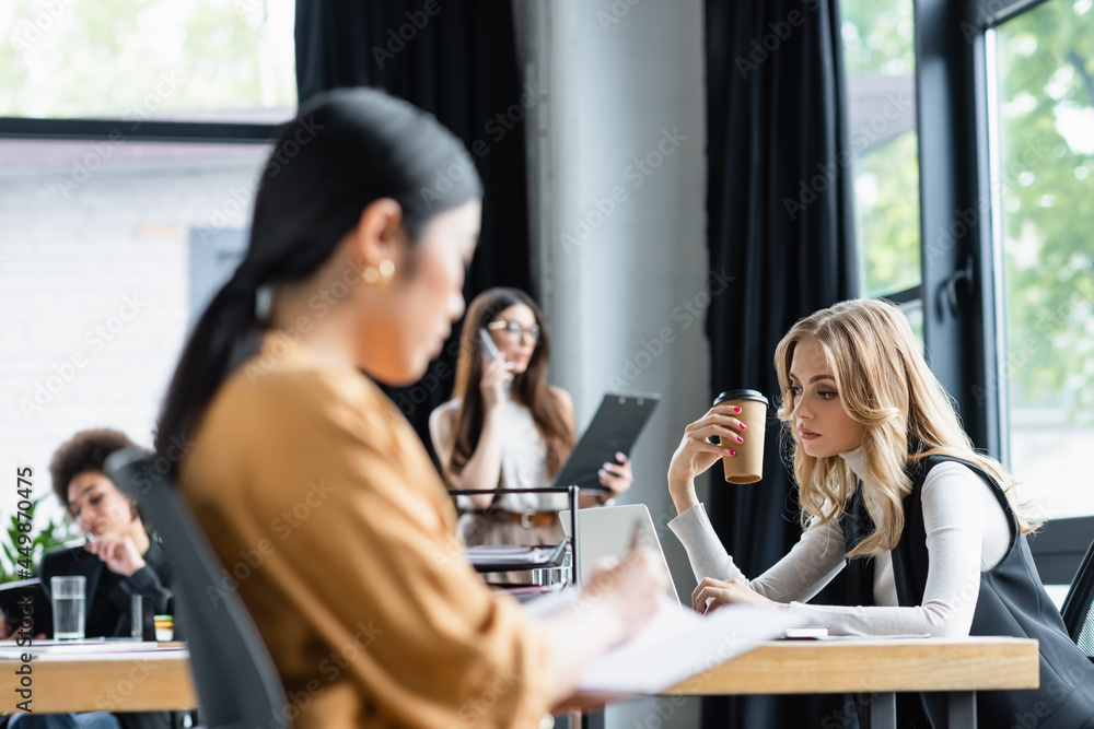blonde businesswoman with coffee to go working on laptop near blurred multiethnic colleagues
