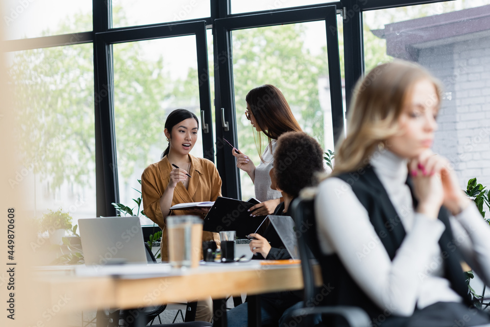 excited asian woman talking to business partner near blurred multiethnic colleagues excited asian woman talking to business partner near blurred multiethnic colleagues
