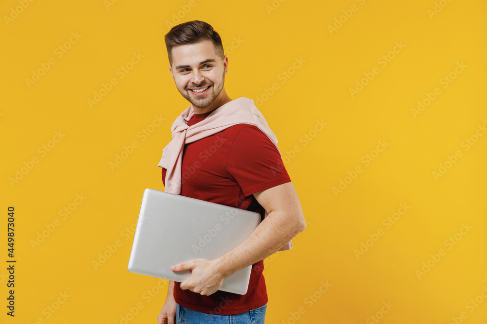 Side view smiling happy young man 20s wear red t-shirt casual clothes use work hold closed laptop pc computer isolated on plain yellow color wall background studio portrait. People lifestyle concept. Side view smiling happy young man 20s wear red t-shirt casual clothes use work hold closed laptop pc computer isolated on plain yellow color wall background studio portrait. People lifestyle concept.