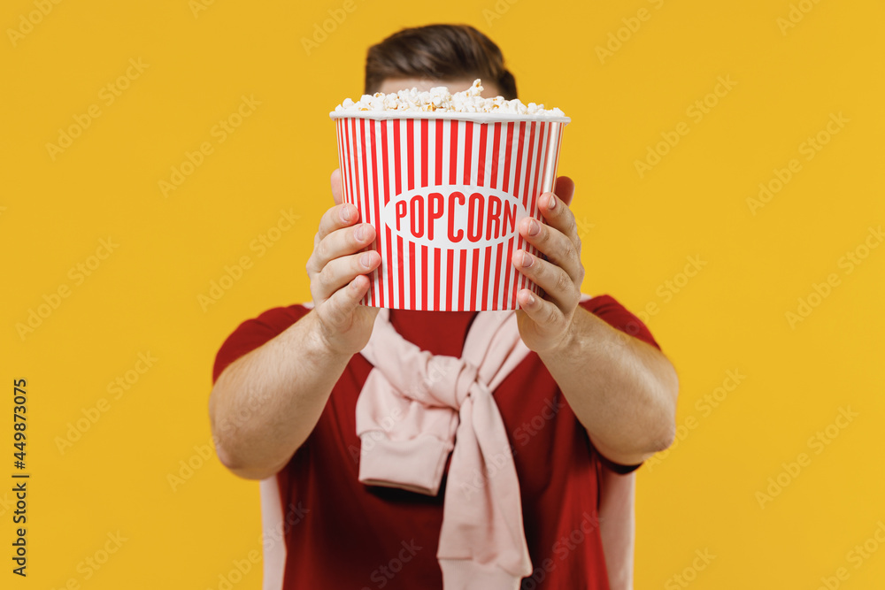 Young caucasian man 20s wearing red t-shirt casual clothes holding showing close up striped takeaway popcorn bucket covering face hiding isolated on plain yellow color wall background studio portrait.
