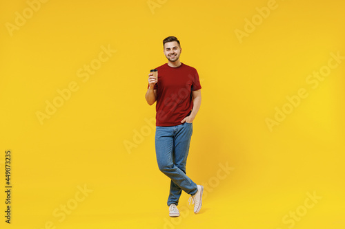 Full length smiling happy fun young man 20s wear red t-shirt casual clothes hold takeaway delivery craft paper brown cup coffee to go isolated on plain yellow color wall background studio portrait