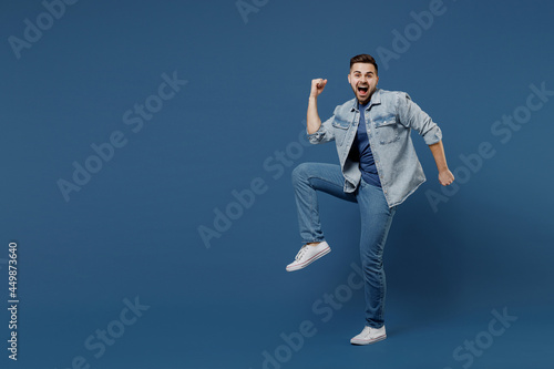 Full size length body laughing beautiful young brunet man 20s mouth open wears denim jacket doing winner gesture celebrating clenching fists say yes isolated on dark blue background studio portrait