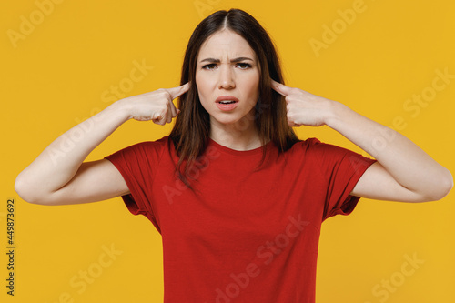 Strict young brunette woman 20s wear basic red t-shirt closed eyes cover ears with hands fingers do not want to listen isolated on yellow background studio portrait. People emotions lifestyle concept