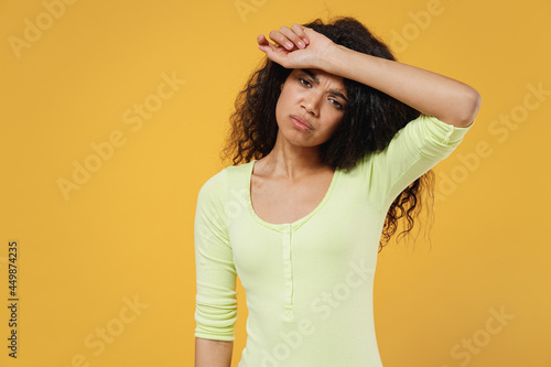 Exhausted fatigued sad african american young brunette woman 20s wears green shirt hand on forehead isolated on yellow background studio portrait. Healthy lifestyle sick ill disease treatment concept.