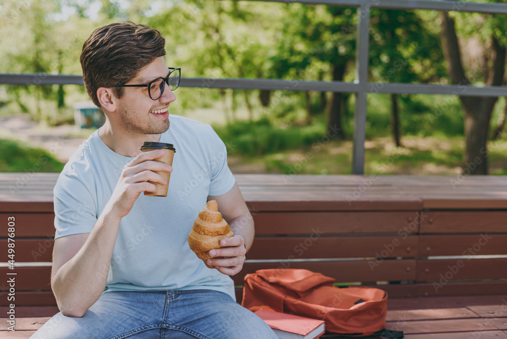 Fototapeta premium Young student man in blue t-shirt eyeglasses backpack sit on bench eat croissant drink coffee have breakfast rest relax in sunshine spring green city park outdoors on nature. Urban leisure concept