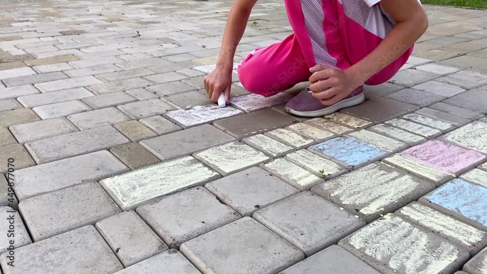 A girl in a white T-shirt and pink pants, squatting, draws with chalk on the paving stones. Side view A girl in a white T-shirt and pink pants, squatting, draws with chalk on the paving stones. Side view