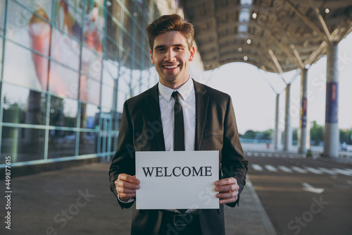 Friendly young satisfied traveler businessman man 20s in black suit stand outside at international airport terminal hold card sign with welcome title text waving hand Air flight business trip concept