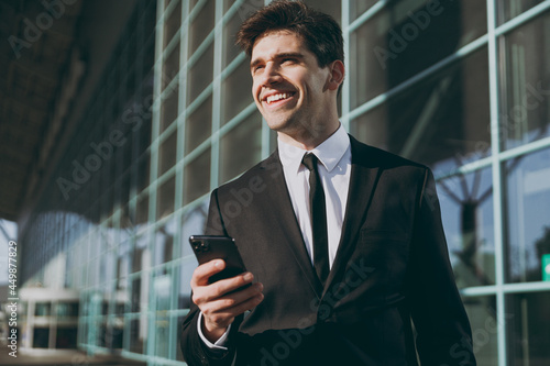 Bottom view smiling young pensive traveler businessman man in black suit stand outside at international airport terminal use mobile cell phone book taxi order hotel Air flight business trip concept.