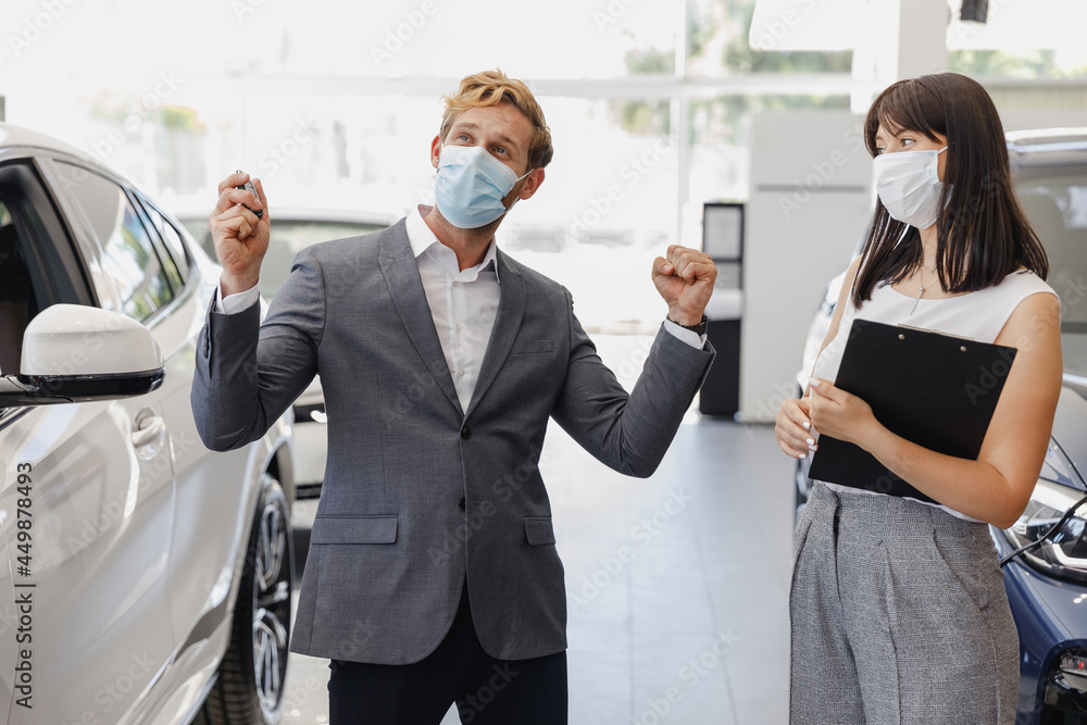 Man winner customer buyer in suit pandemic mask choose auto want buy new car automobile consult with salesman hold keys clench fist in showroom salon dealership store motor show indoor Sale concept. Man winner customer buyer in suit pandemic mask choose auto want buy new car automobile consult with salesman hold keys clench fist in showroom salon dealership store motor show indoor Sale concept.