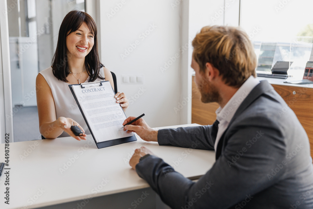 Friendly female woman salesman wearing shite shirt give keys fob showing contract for signature of customer client male buyer in grey suit sit at office in salon dealership store indoor Sale concept Friendly female woman salesman wearing shite shirt give keys fob showing contract for signature of customer client male buyer in grey suit sit at office in salon dealership store indoor Sale concept
