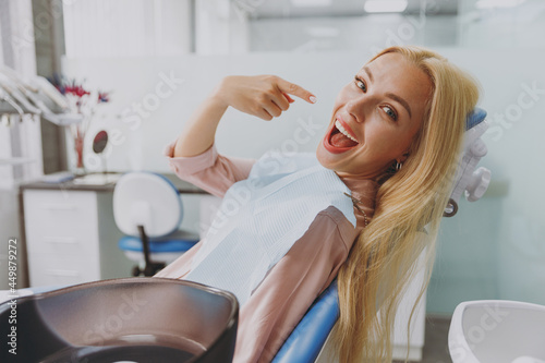 Young smiling happy woman covered by napkin point index finger on healthy toothy smile sit at dental office chair indoor light modern cabinet wait for stomatologist Healthcare oral enamel treatment