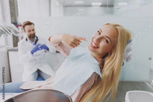 Young smiling happy woman 20s with dental napkin point finger on mouth sit at dentist office chair indoor light cabinet near stomatologist wait for oral procedure Healthcare caries enamel treatment.