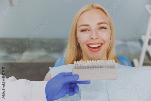 Close up young smiling happy fun woman 20s choosing veneers enamel color palette sit at dentist office chair indoor light modern cabinet near stomatologist Healthcare oral enamel aesthetic treatment.