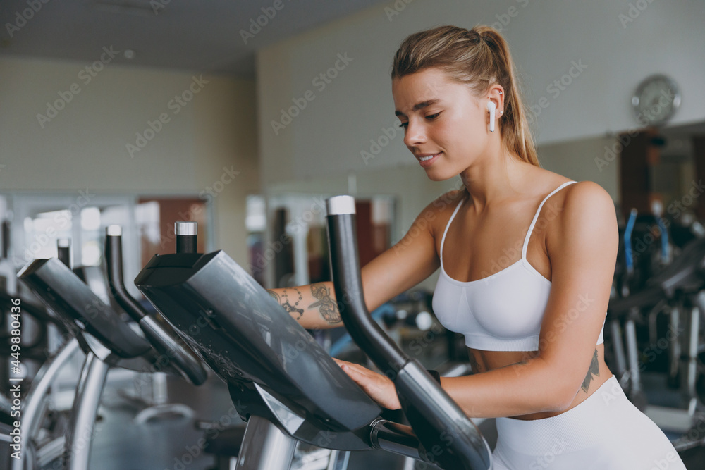 Side view minded concentrated serious young strong sporty athletic sportswoman woman in white sportswear earphones listen music warm up training use exercise bike in gym indoor Workout sport concept Side view minded concentrated serious young strong sporty athletic sportswoman woman in white sportswear earphones listen music warm up training use exercise bike in gym indoor Workout sport concept