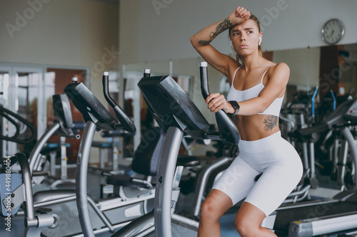 Young tired strong sporty athletic sportswoman caucasian woman in white sportswear earphones listen music put hand forehead sweat warm up train use exercise bike in gym indoor Workout sport concept.