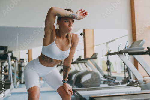 Photos Young tired strong sporty athletic sportswoman woman in white sportswear warm up train near treadmill stand leaning on knees put hand on head breathing in gym indoor Workout sport motivation concept